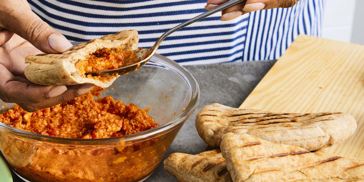 Person in a striped apron standing behind a grey table filling a pita pocket with romesco sauce over a glass bowl of romesco sauce