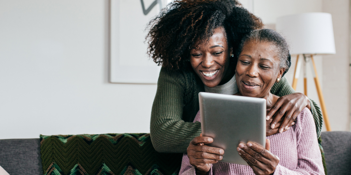 Senior woman on her tablet in the living room with a young female caregiver behind her.