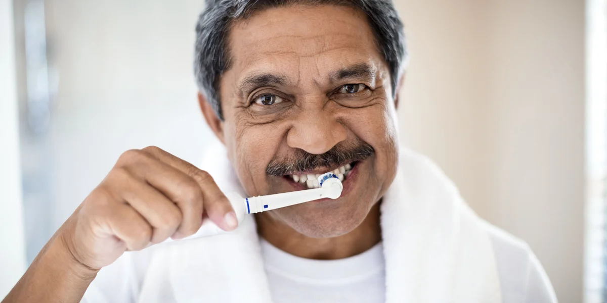Man in a white shirt with a white towel around his neck brushing his teeth with an electric toothbrush