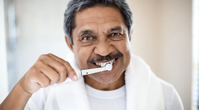 Man in a white shirt with a white towel around his neck brushing his teeth with an electric toothbrush