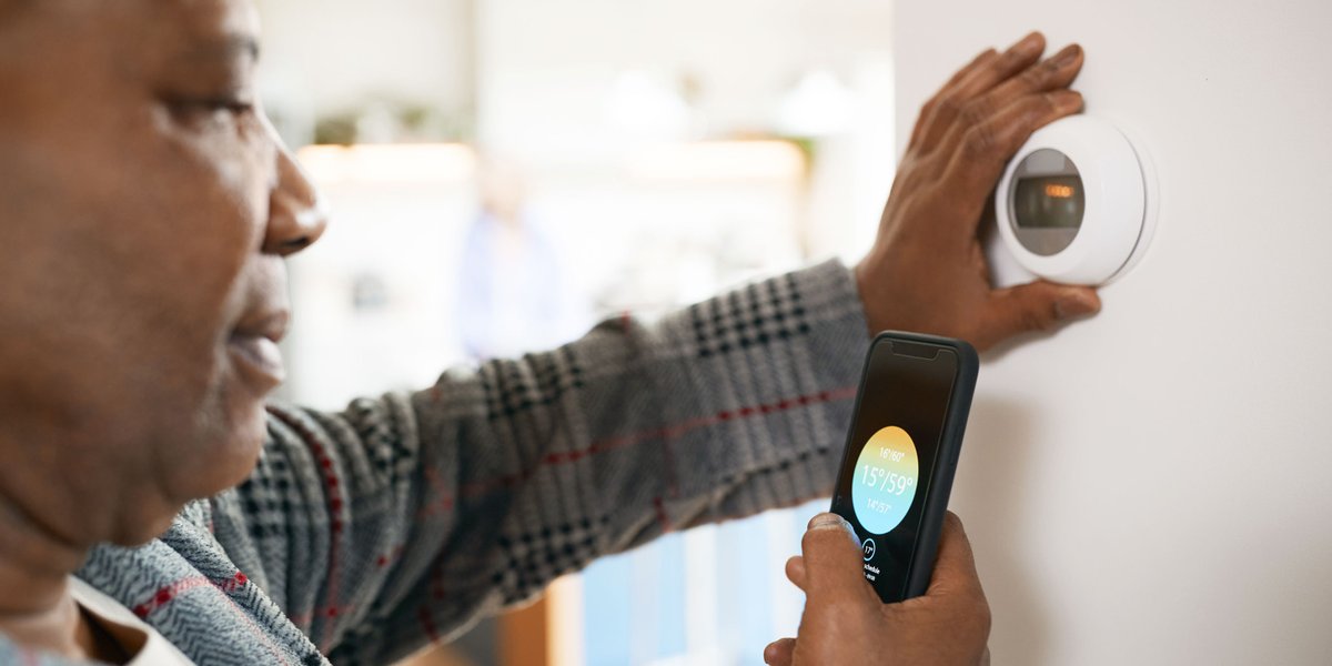 Woman standing in front of her smart thermostat while using her phone to adjust the temperature
