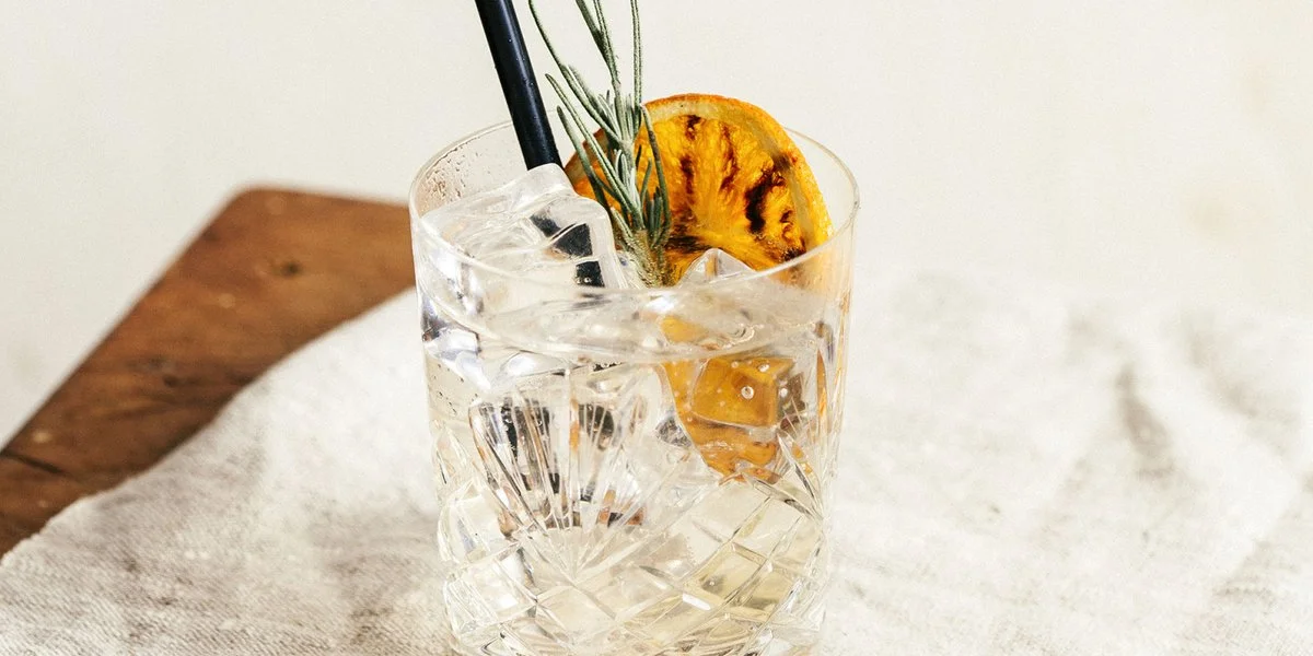 Glass of orange tonic with a sprig of rosemary and burnt orange garnish placed on a white table cloth