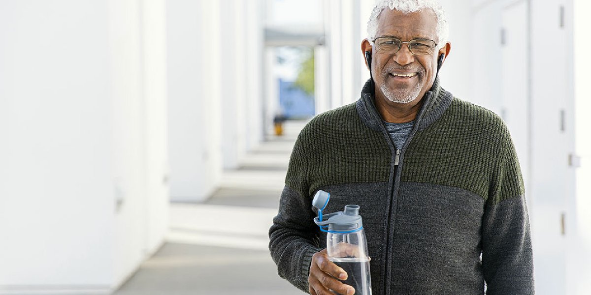 Man in a black sweater with earbuds in, holding a water bottle while standing in a hallway