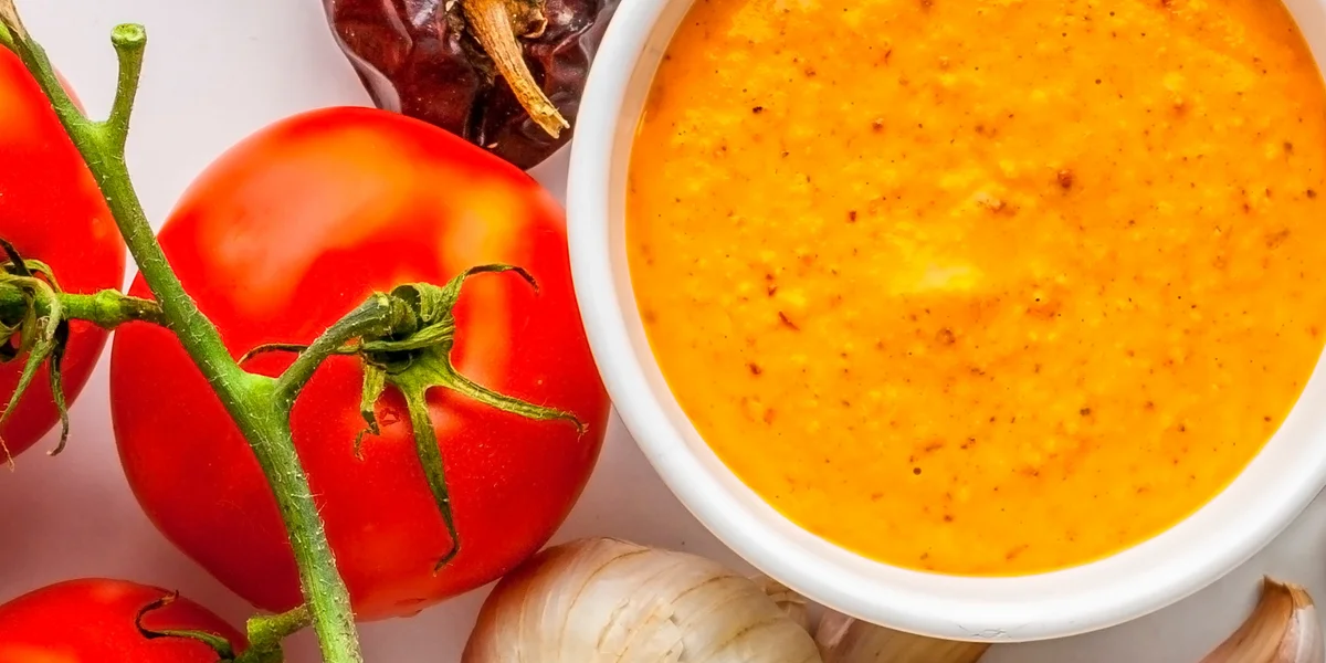Bowl of romesco sauce surrounded by fresh tomatoes, garlic cloves, and roasted bell peppers.