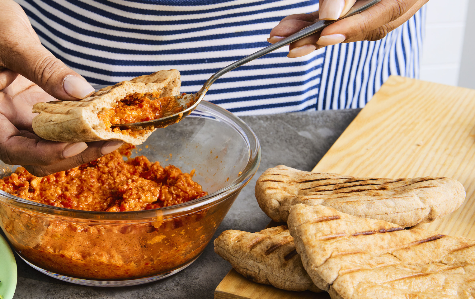 Person in a striped apron standing behind a grey table filling a pita pocket with romesco sauce over a glass bowl of romesco sauce