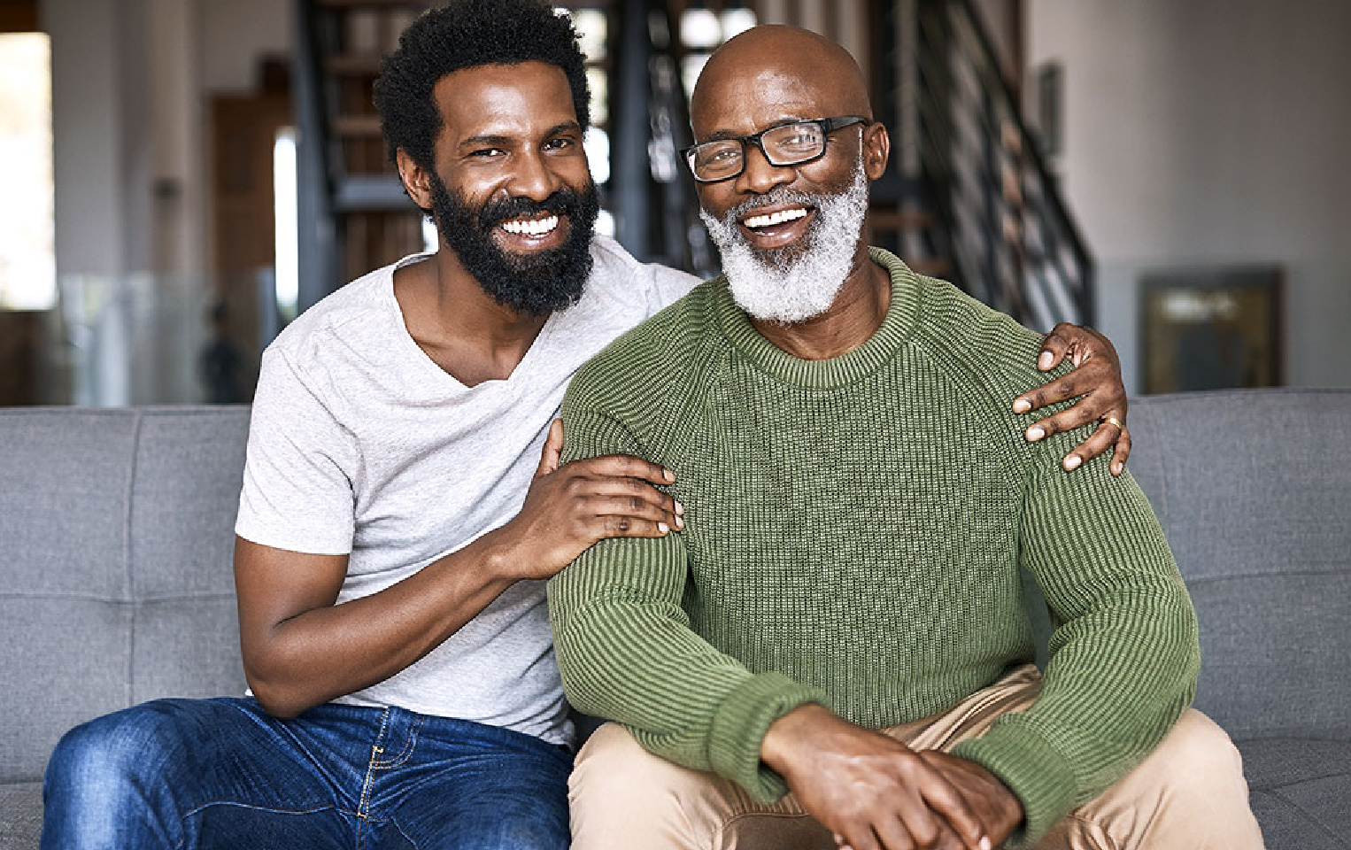 Man in a grey shirt smiling and sitting on the couch next to his father in a green sweater