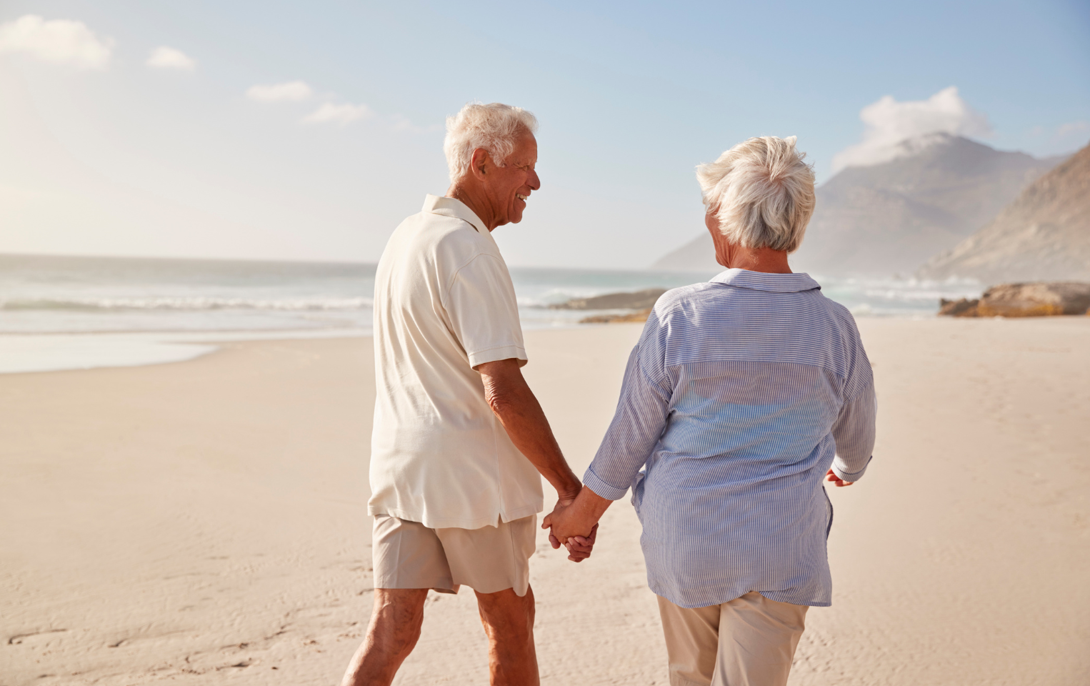 Elderly couple smiling and holding hands while walking on an empty beach.