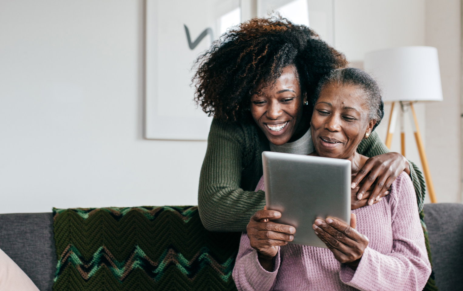 Senior woman on her tablet in the living room with a young female caregiver behind her.