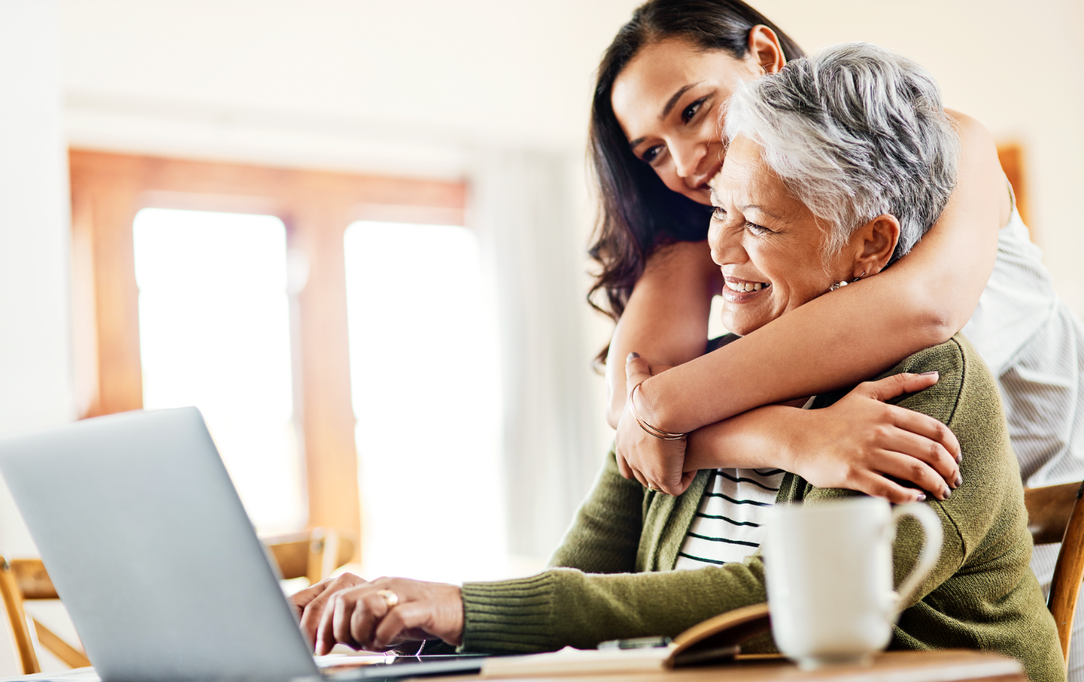 Older woman smiling at a table on her laptop with a younger woman hugging her.