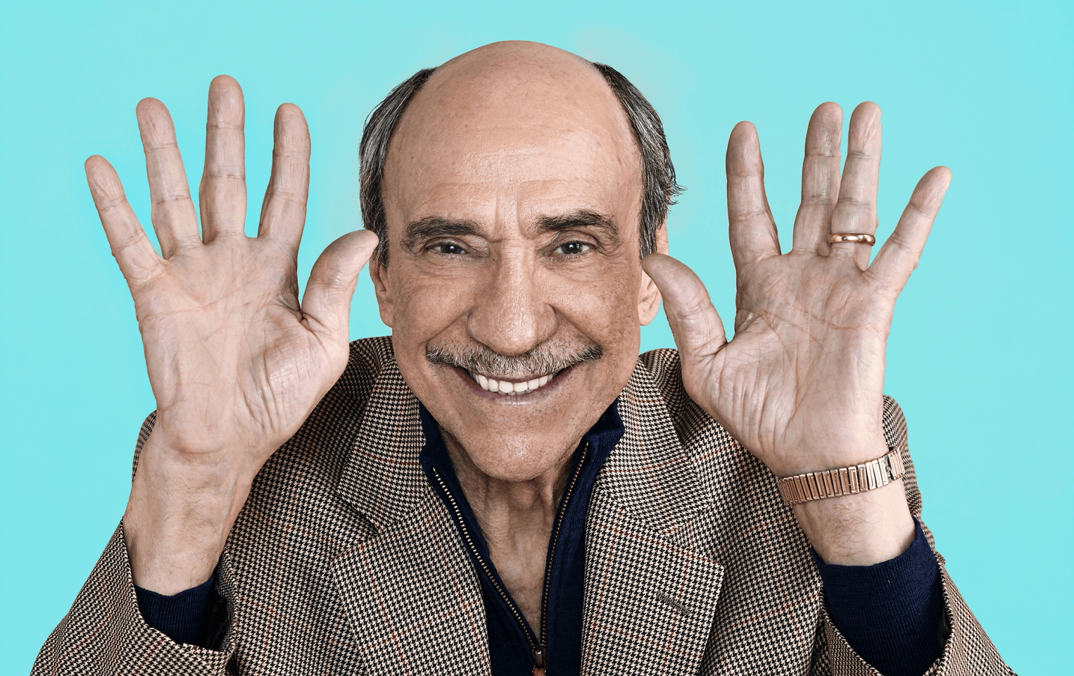 F. Murray Abraham standing against a blue background holding his hands up near his face