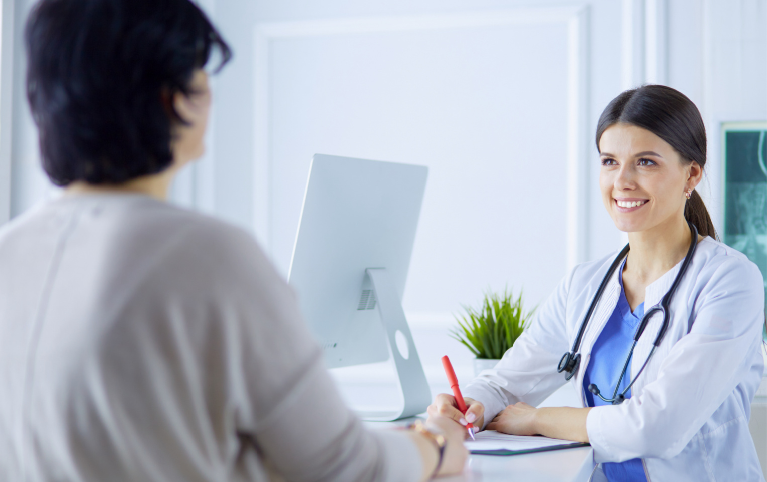 Doctor in a white lab coat and blue scrubs speaking to a patient in a grey sweater at a doctor's office while writing down notes