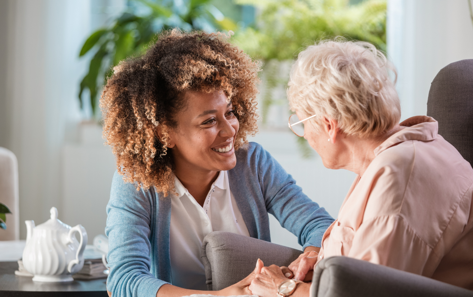 Smiling female caregiver in a blue cardigan holding hands and talking with senior woman in living room