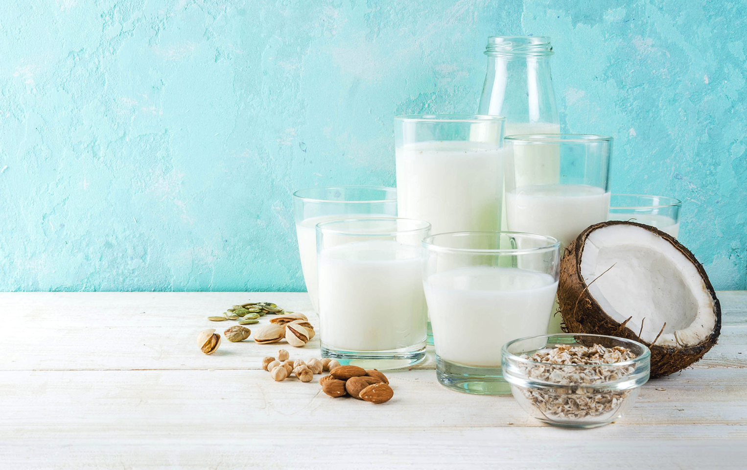 Variety of alternative milks in glasses on a white table in front of a blue background with small bowls of soy, almond, flax, oats, and half of a coconut.