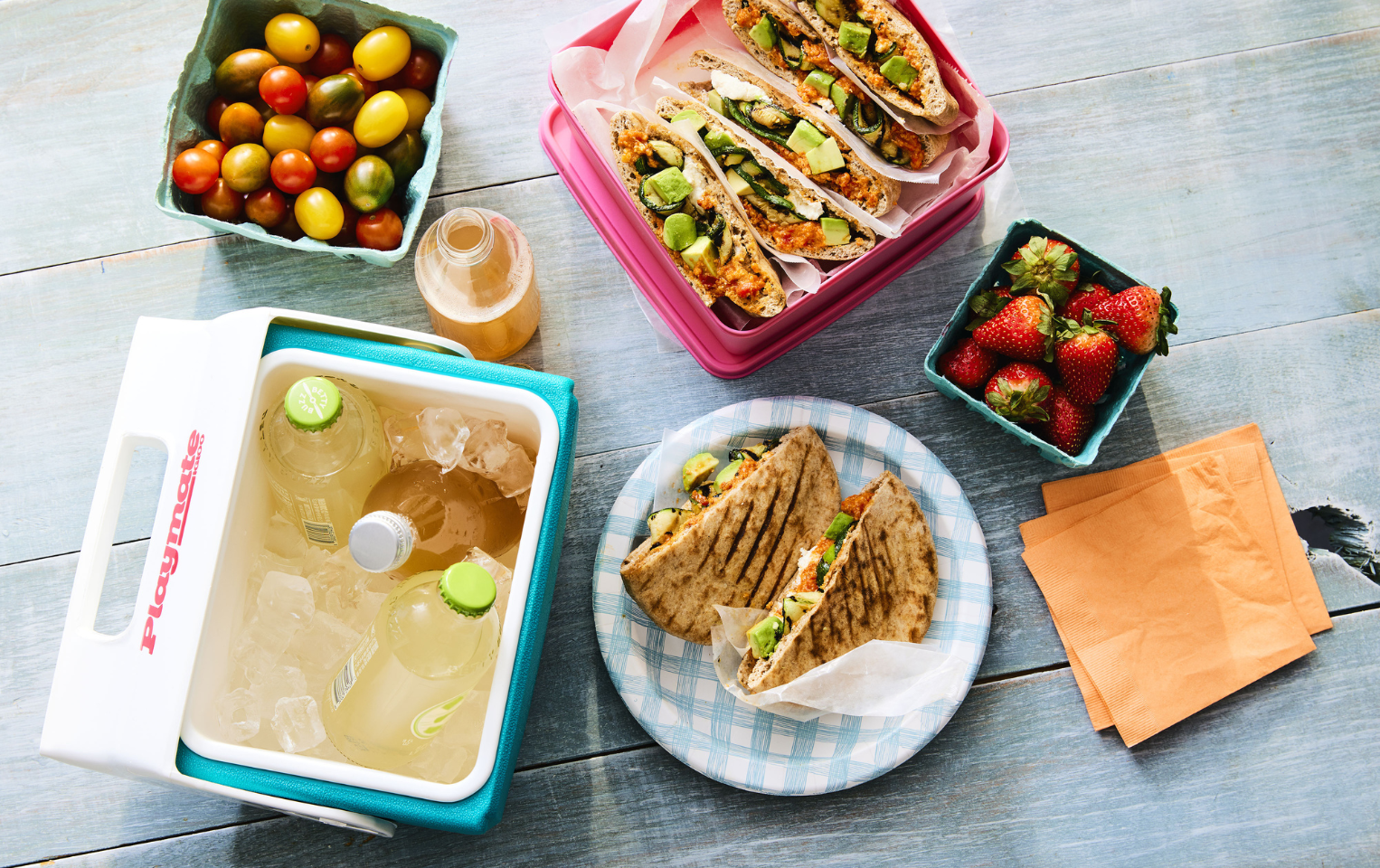 A plate and container with zucchini and romesco sandwich surrounded by fresh strawberries, tomatoes, and a cooler with refreshing beverages on a wooden grey table