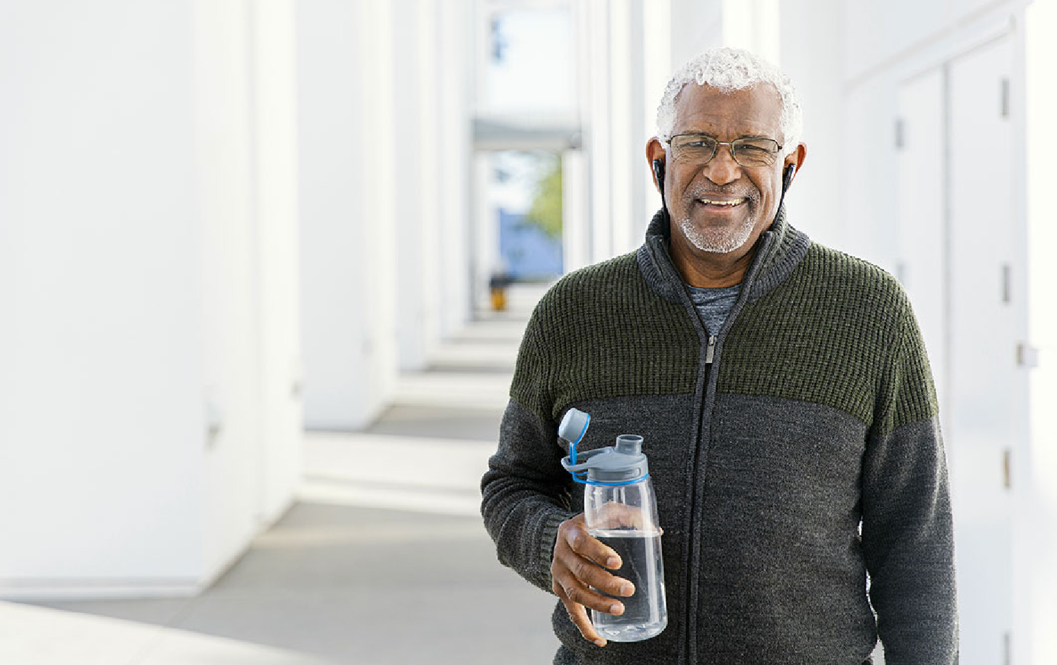 Man in a black sweater with earbuds in, holding a water bottle while standing in a hallway