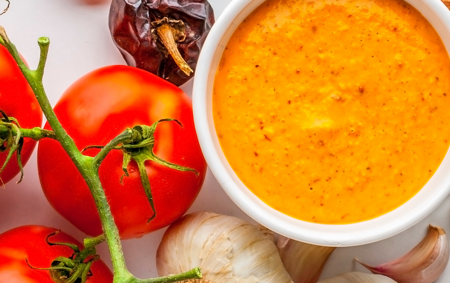 Bowl of romesco sauce surrounded by fresh tomatoes, garlic cloves, and roasted bell peppers.