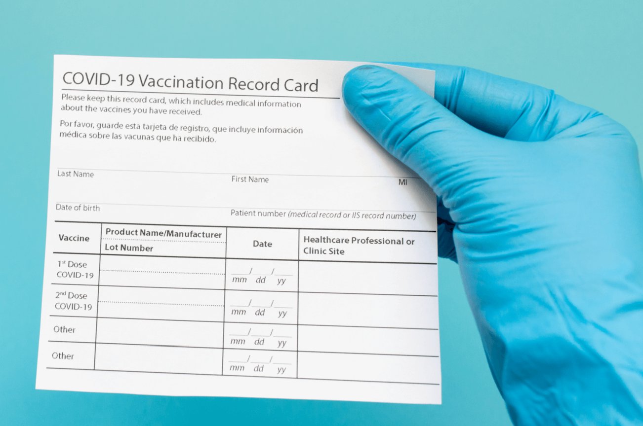 hand with a blue medical glove holding a COVID-19 vaccination record card up against a blue background